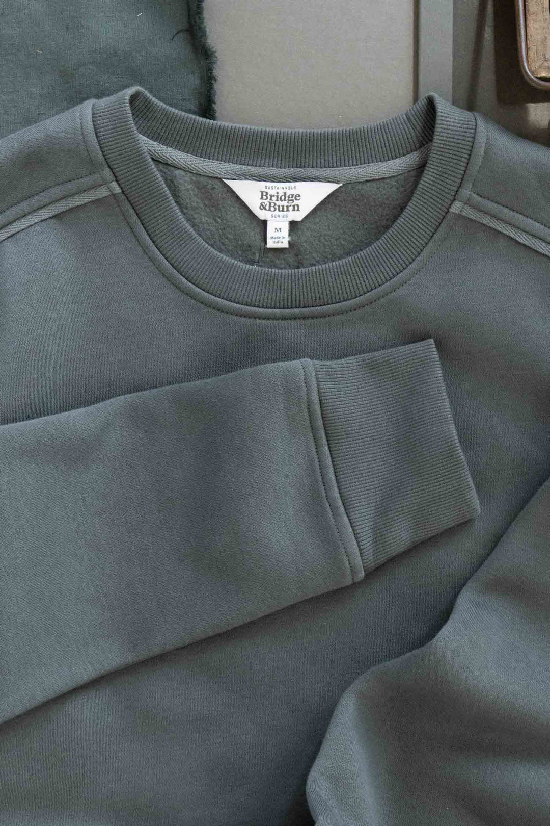 A close-up of the Bailey Crew Sweatshirt in Slate by Bridge & Burn, crafted from soft, sustainable materials. The folded crewneck showcases its label and is laid flat on a surface.