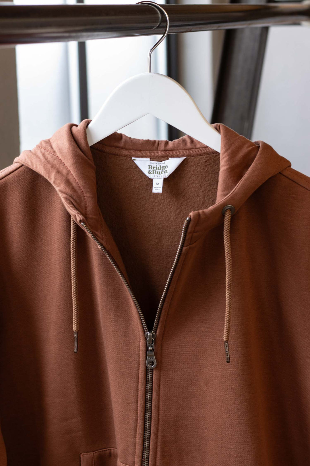 The Bridge & Burn Mason Hoodie in Burnt Umber hangs on a white hanger, showcasing its front zipper, drawstrings, and signature tag against a softly blurred neutral background.