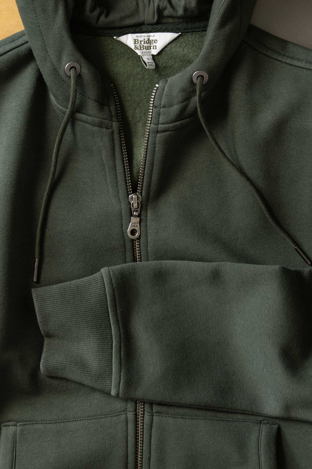 The Bridge & Burn Mason Hoodie in Forest Night, a sustainable zip-front sweatshirt with drawstrings, is shown laid flat with a partially open zipper and one sleeve folded over the body. The inner label shows the brand name.