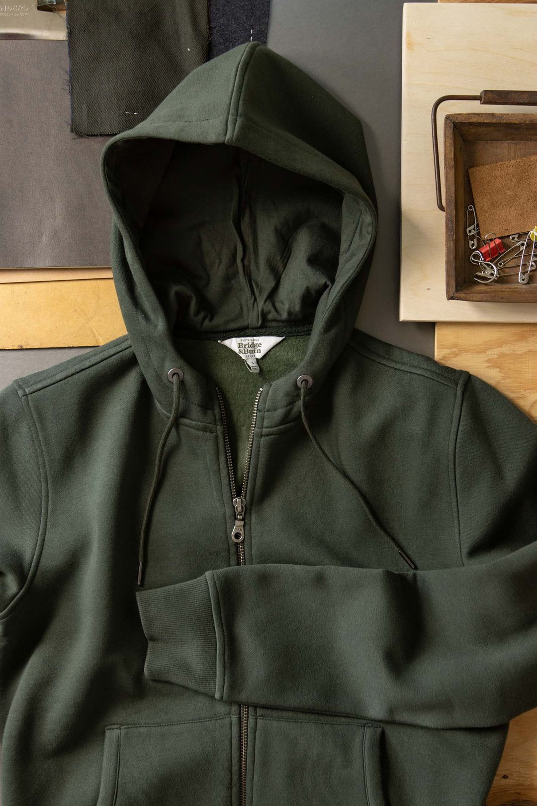 The Bridge & Burn Mason Hoodie in Forest Night, made from organic cotton, is laid flat on a table with fabric swatches and a wooden tray holding keys and paper clips nearby.