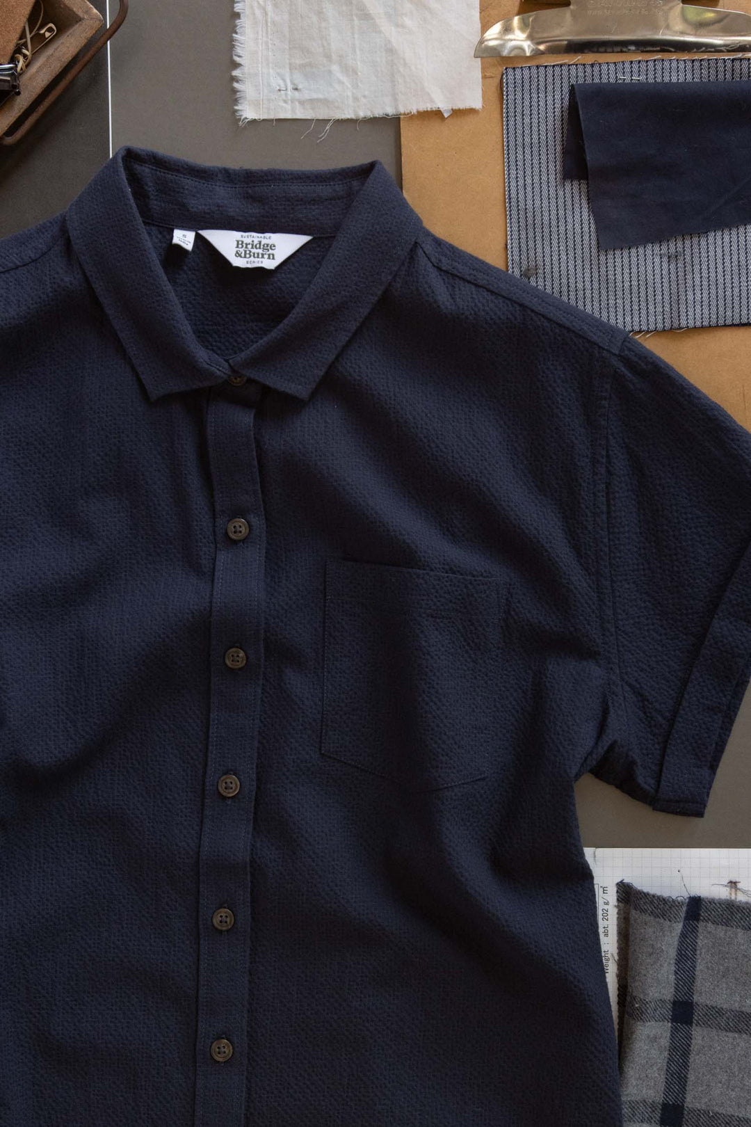 The Bridge & Burn Bea Shirt in Navy Seersucker is a short-sleeve button-down with a chest pocket, shown laid flat and surrounded by fabric swatches, a brown folder, and sewing materials.