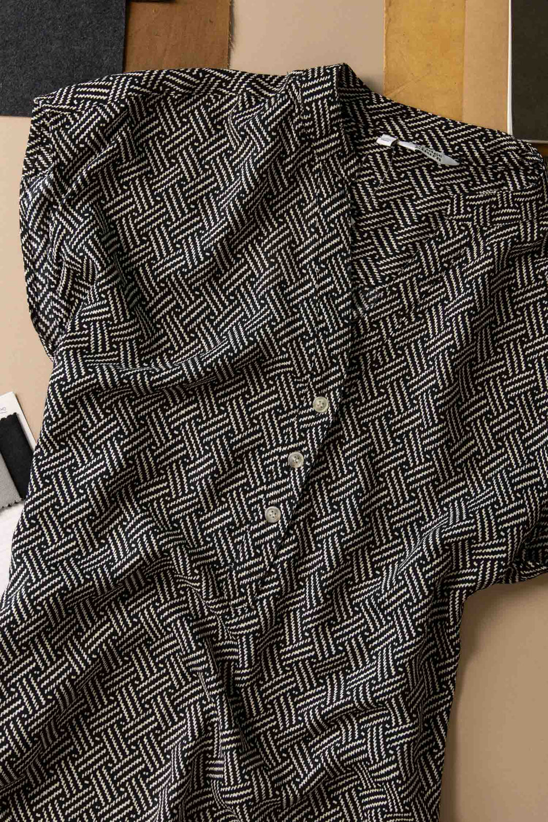 A close-up of the Bridge & Burn Ladd Shirt in Black Geo, a short sleeve top with a black and white geometric pattern, folded on a neutral surface with fabric swatches nearby.