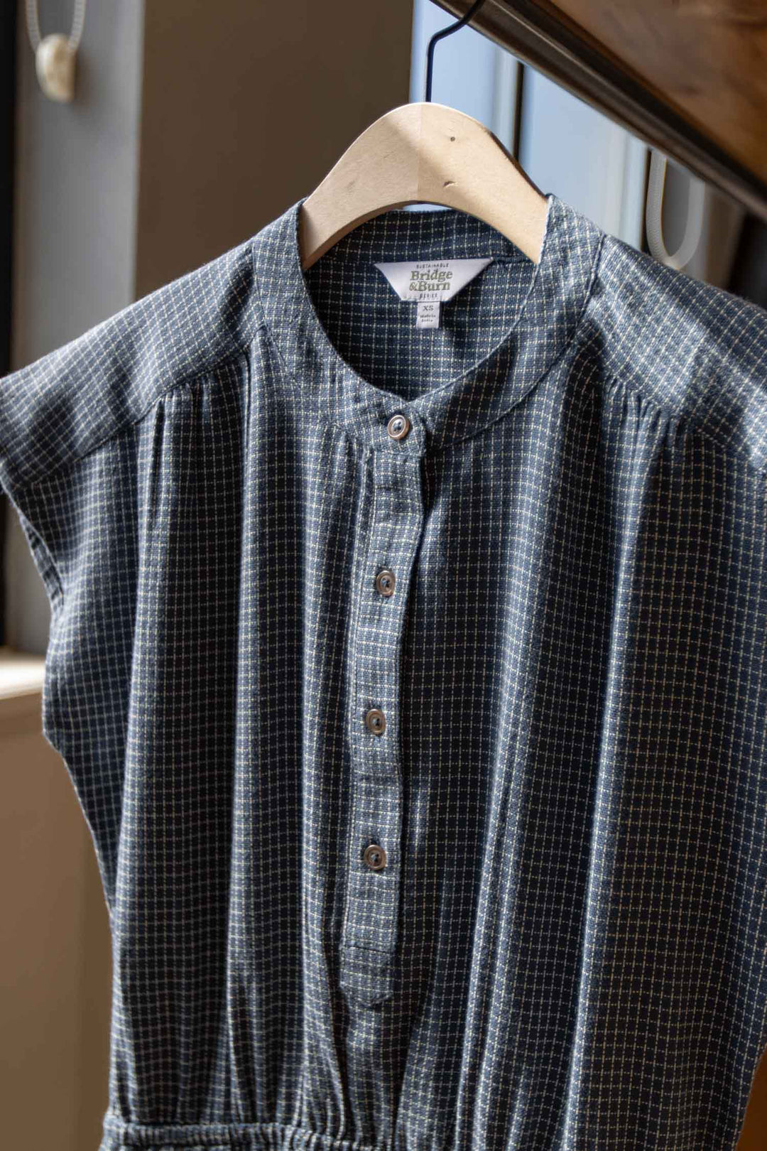 The Lorane Dress in Navy Grid by Bridge & Burn, an A-line cotton dress with short sleeves, hangs on a wooden hanger near a window as sunlight softly illuminates its classic blue and white check pattern.