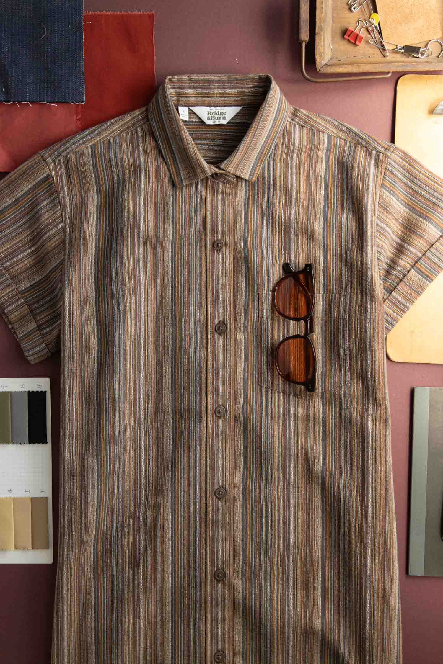 The Bridge & Burn Loren Shirt Dress in Multistripe, from the Sustainable Series Collection, is laid flat with brown sunglasses in the chest pocket and surrounded by fabric swatches and design materials.