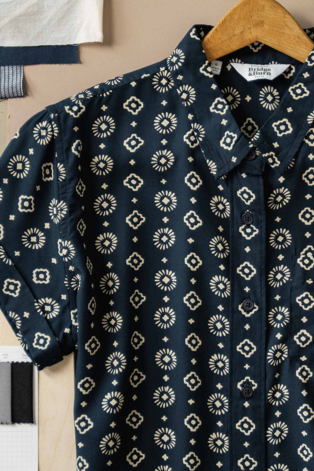 The Bridge & Burn Bea Shirt in Midnight Sundance is a dark blue, short-sleeve button down with a tailored fit and cream geometric floral pattern. Shown on a wooden hanger against beige with visible fabric swatches in the corners.