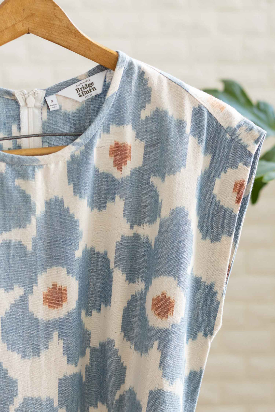 The Eloise Dress in Polar Blossom by Bridge & Burn features a blue and brown abstract floral print on cream fabric, sleeveless with an A-line silhouette. Displayed on a wooden hanger against a light brick wall with a green plant nearby.
