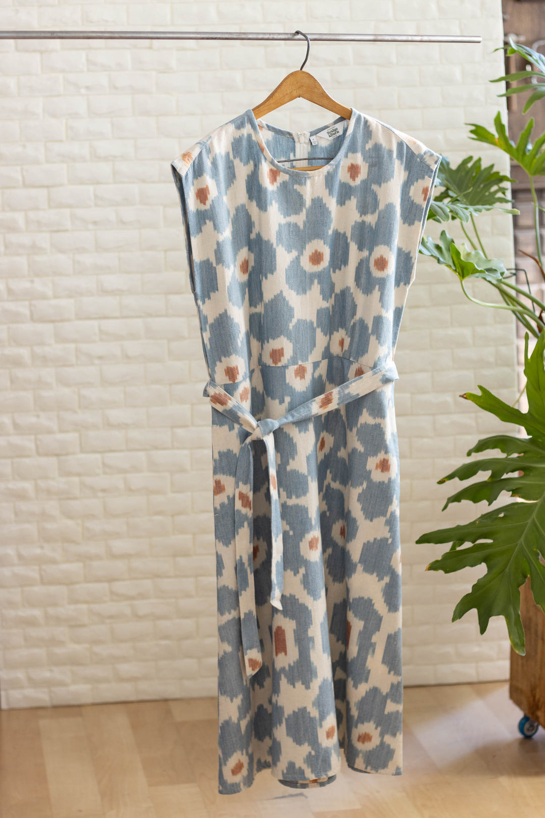 The Eloise Dress in Polar Blossom by Bridge & Burn, featuring a blue and white abstract floral print and a matching waist belt, hangs on a wooden hanger against white tile, with a green leafy plant nearby.