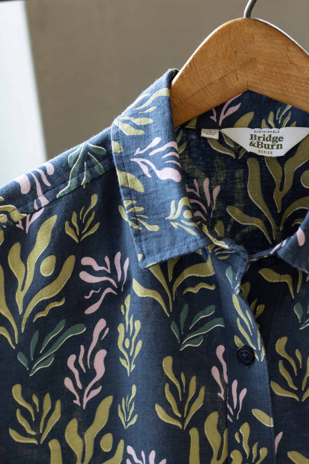 A close-up of the Greer Cropped Shirt in Spring Kelp by Bridge & Burn, featuring a blue cotton-linen blend with a green and pink leafy pattern, hanging on a wooden hanger.