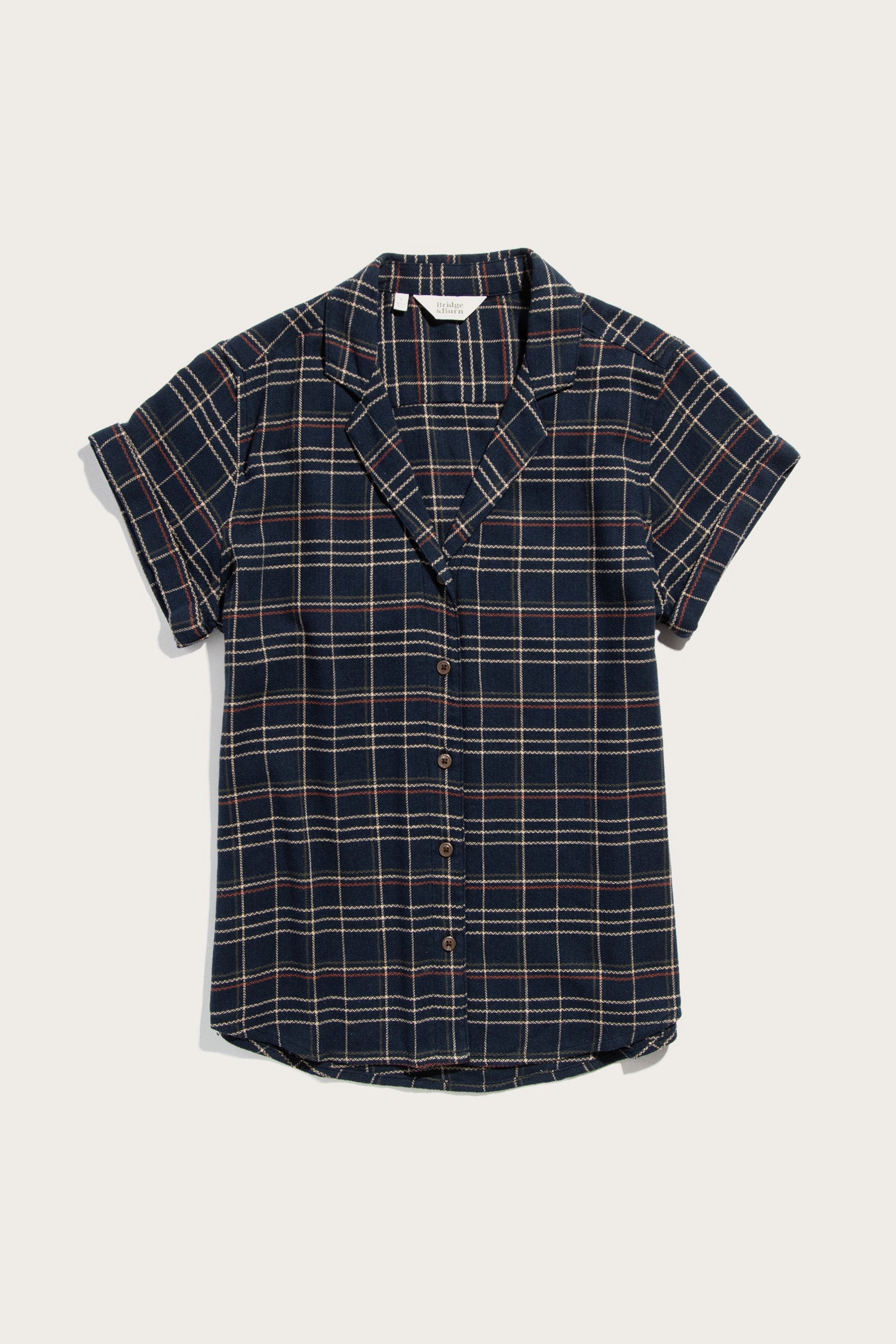 The Bridge & Burn Innes Shirt in Dusk Herringbone Plaid is a relaxed fit, short-sleeve button-up made from recycled cotton, featuring a navy and white plaid, notched collar, and cuffed sleeves. Laid flat on a white background.
