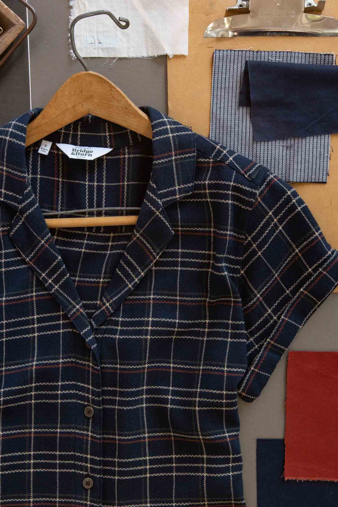 The Bridge & Burn Innes Shirt in Dusk Herringbone Plaid—a relaxed fit, short-sleeve button-up—features a navy, red, and white pattern. Made from recycled cotton, it's displayed on a wooden hanger with swatches and papers in the background.