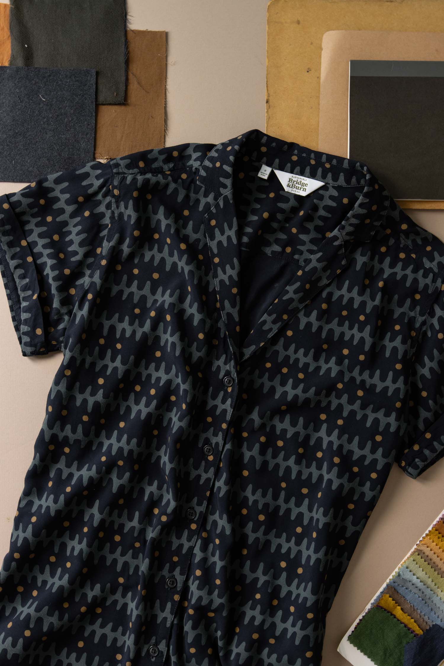 The Innes Shirt / Fishbone Ink by Bridge & Burn is a relaxed-fit, short-sleeve button-up with a lapel collar and dark blue zigzag dotted pattern, displayed on beige among fabric swatches and paper samples.