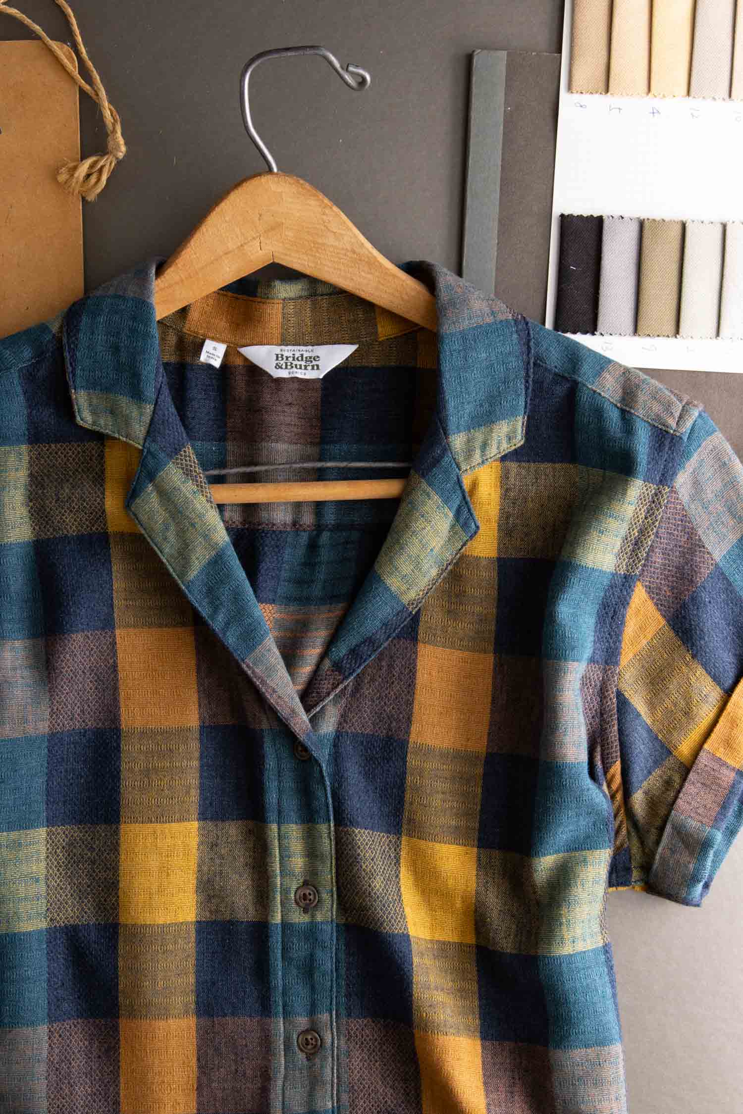 The Bridge & Burn Innes Shirt in Moonbow Plaid, made from recycled cotton, features a relaxed fit and blue, yellow, and brown checks. Shown on a wooden hanger with fabric swatches and crafting materials on a dark surface.