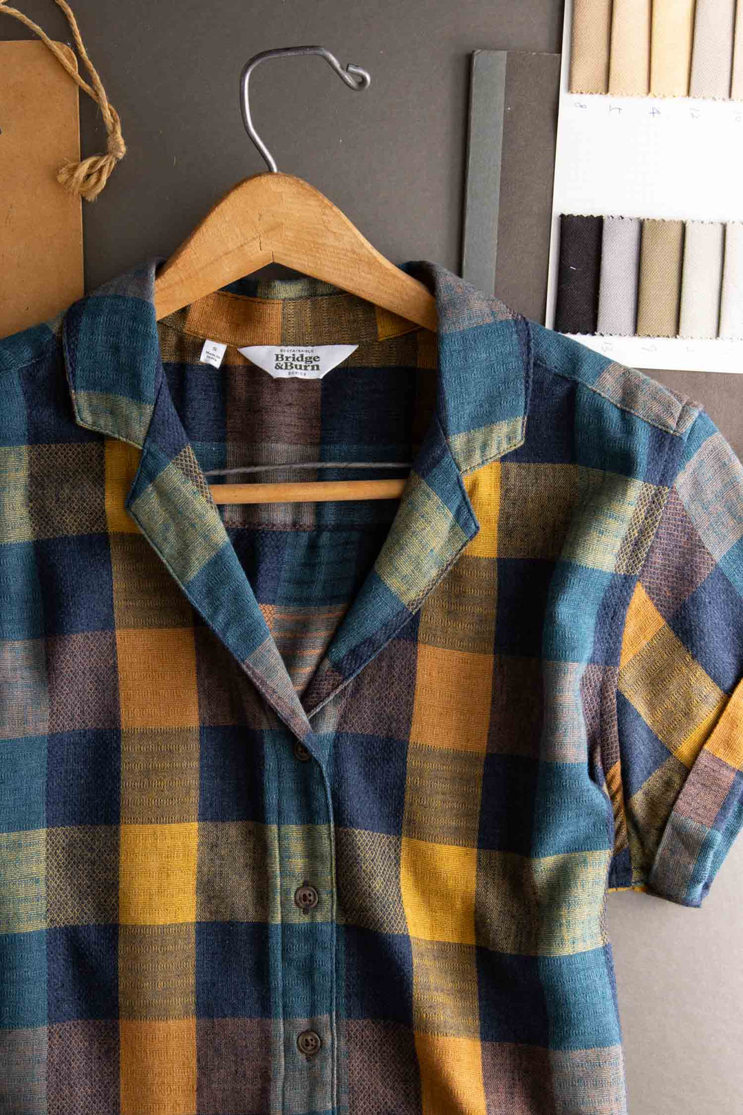 The Bridge & Burn Innes Shirt in Moonbow Plaid, made from recycled cotton, features a relaxed fit and blue, yellow, and brown checks. Shown on a wooden hanger with fabric swatches and crafting materials on a dark surface.