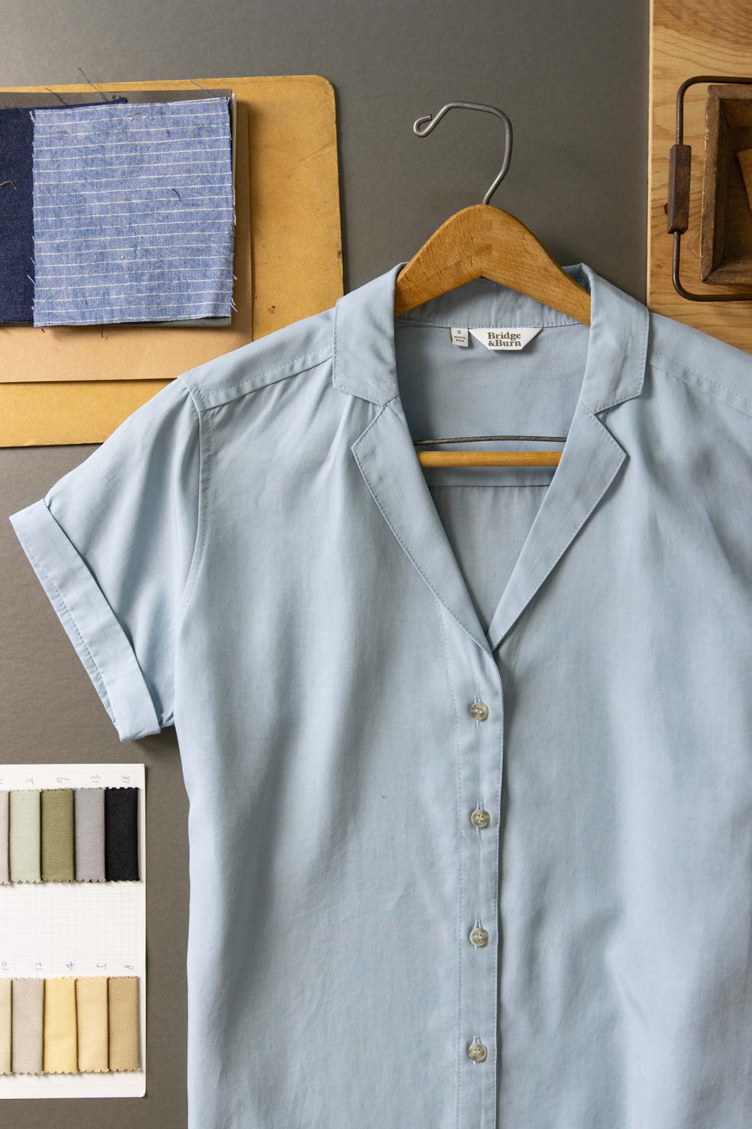 The Bridge & Burn Innes Shirt in Sky Blue, a relaxed fit button-up, is displayed on a wooden hanger over a gray surface, surrounded by fabric swatches and a sewing pattern.