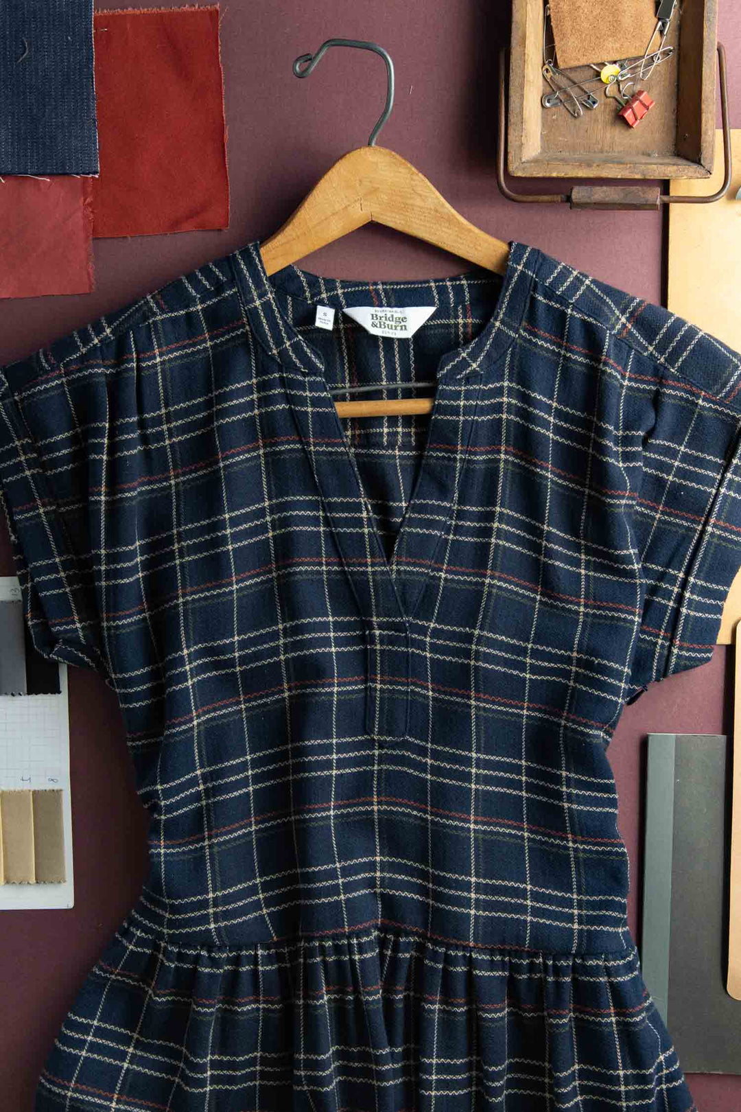 The Bridge & Burn Ira Dress in Dusk Herringbone Plaid, a short-sleeved navy pullover made from recycled cotton with white and red plaid, is displayed on a wooden hanger against a purple wall amid fabric swatches and sewing supplies.