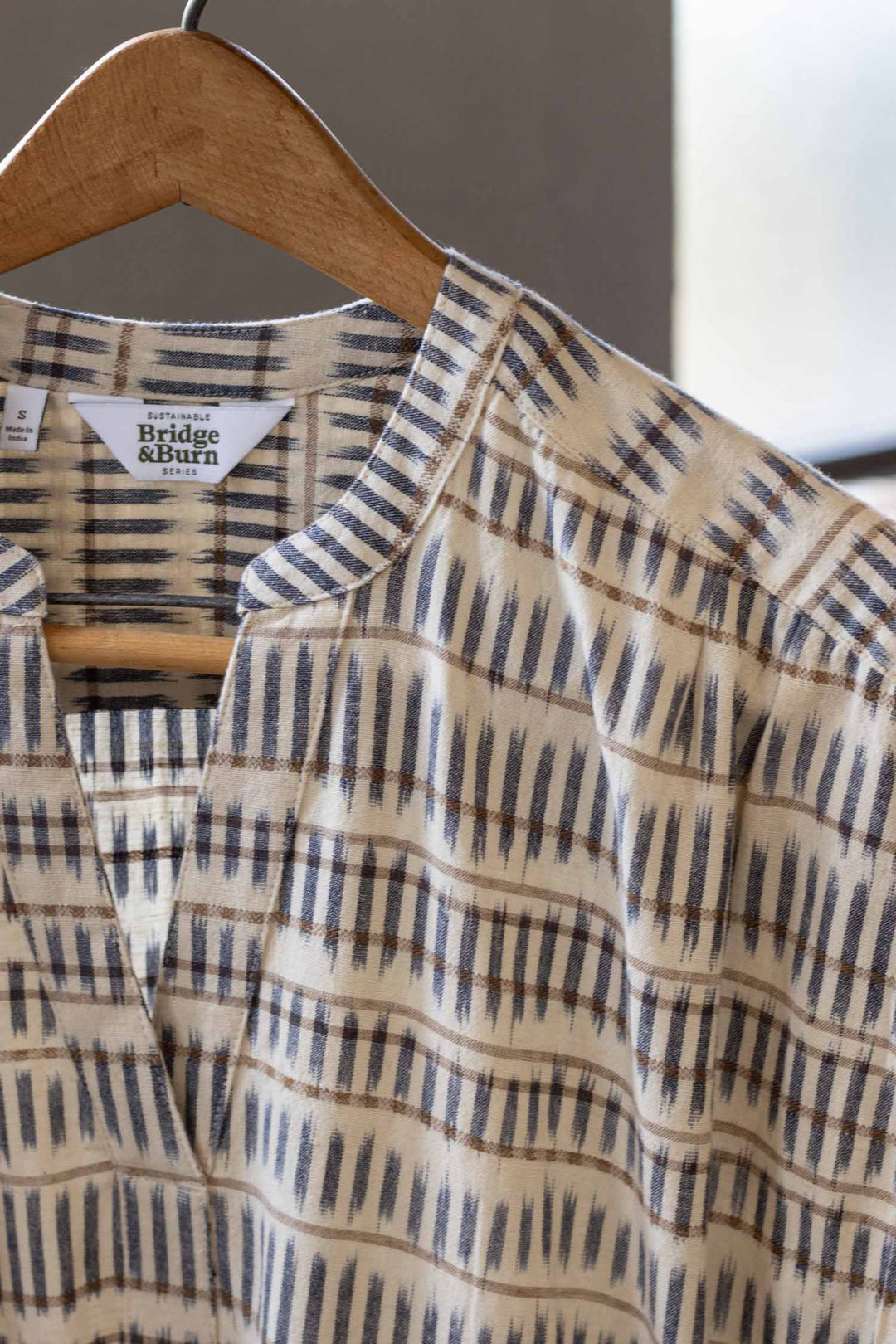 The Bridge & Burn Ira Dress in Mushroom Ikat Stripe, featuring cream fabric with blue and brown geometric patterns, hangs on a wooden hanger with sunlight streaming through a window in the background.