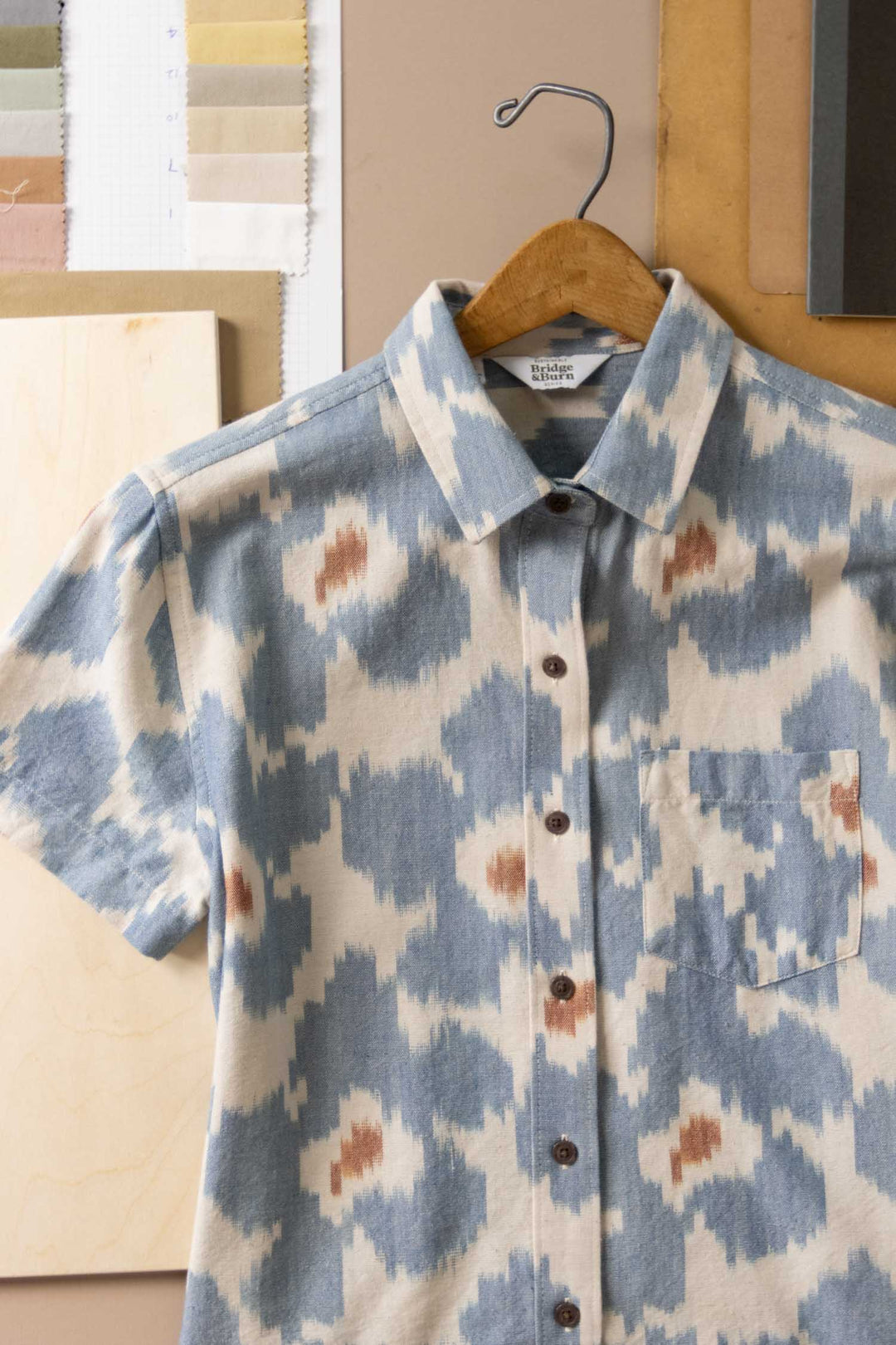 The Bridge & Burn Lana Shirt in Polar Blossom, a relaxed fit short sleeve button-up featuring a blue and brown abstract ikat pattern, hangs on a wooden hanger against a neutral background with fabric swatches and papers.