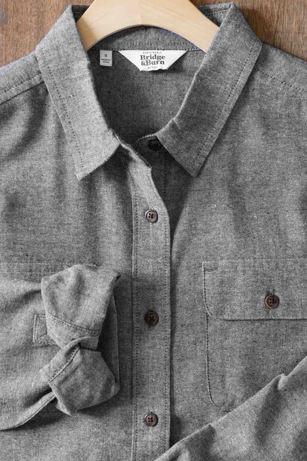 The Lenon Shirt in Iron Chambray by Bridge & Burn is shown on a wooden hanger, with one sleeve partially rolled up and set on a wooden surface. This relaxed fit gray shirt features a front pocket.