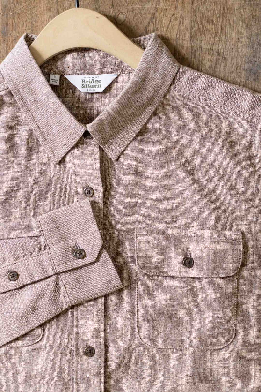The Bridge & Burn Lenon Shirt in Rust Chambray is a light brown, long-sleeve relaxed fit shirt with a single chest pocket, crafted from recycled cotton with a subtly textured look, shown on a wooden hanger against a wood background.