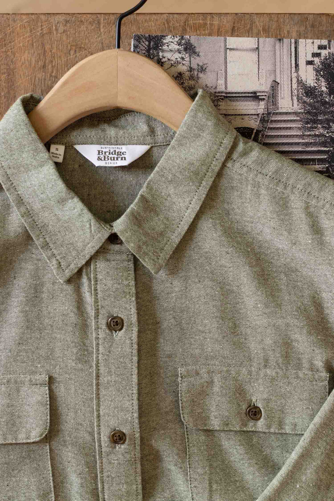 A close-up of the Bridge & Burn Lenon Shirt in Sage Chambray—made from recycled cotton—displays its collar, chest pockets, and buttons on a wooden hanger, with a black-and-white photo partially visible in the background.