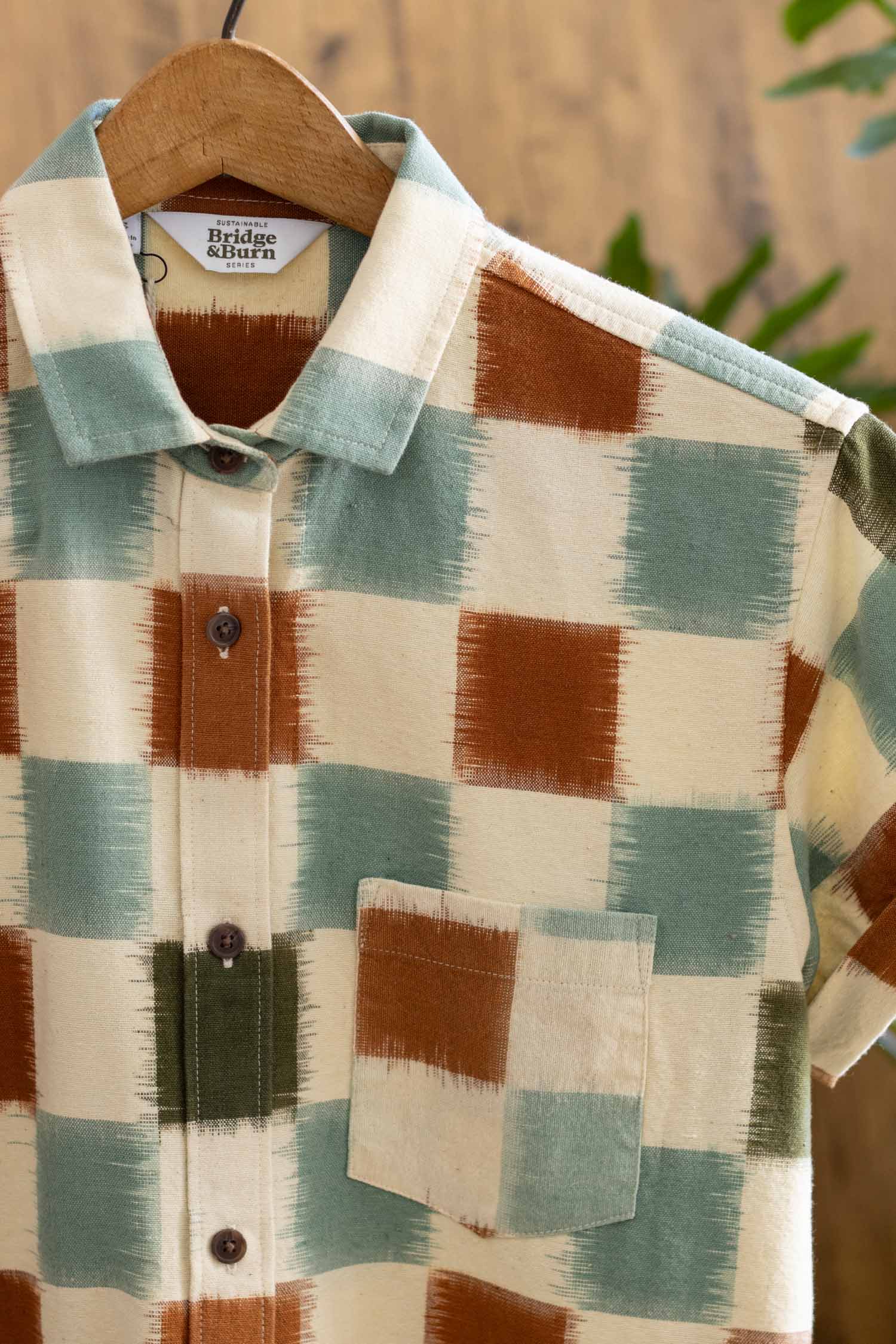 The Loren Shirt Dress / Mod Checker by Bridge & Burn is a short-sleeve, 100% cotton dress with a bold checkered pattern in cream, rust orange, sage green, and olive. Featuring a front pocket, it's shown on a wooden hanger against a blurred backdrop.