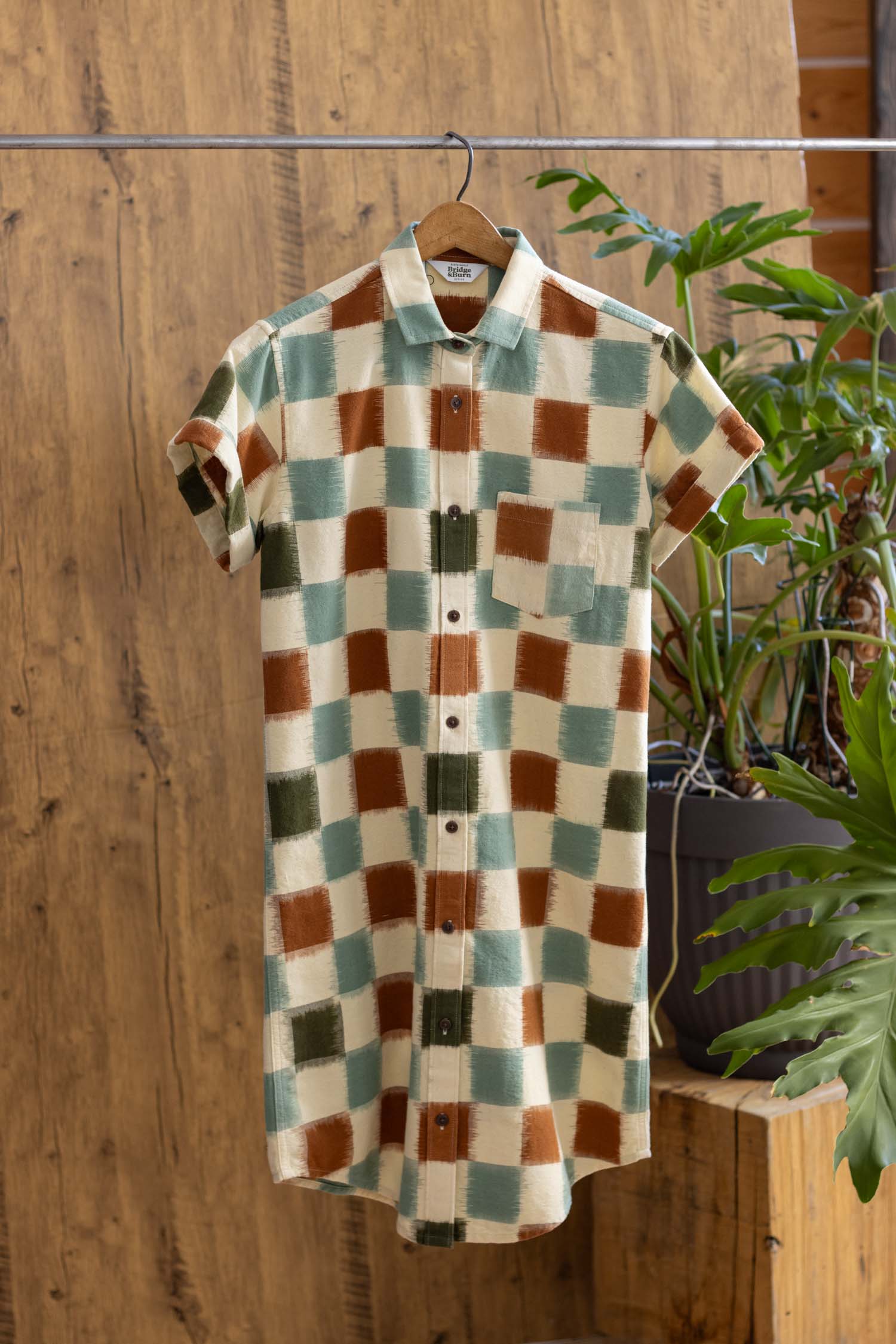 The Loren Shirt Dress / Mod Checker by Bridge & Burn, made from 100% cotton with a green, brown, tan, and cream checkered pattern, hangs on a wooden hanger amid green plants against a wooden backdrop.