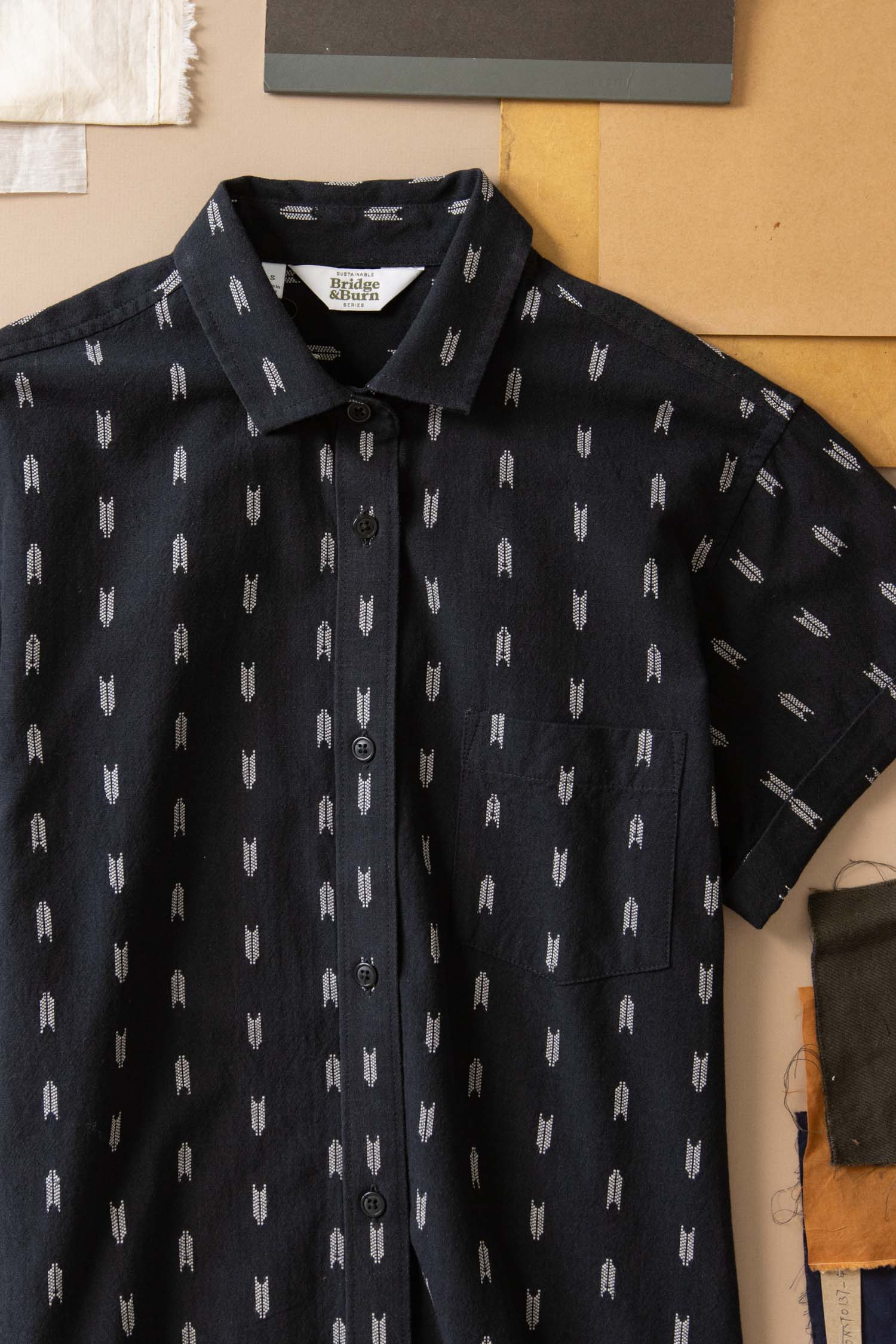 The Loren Shirt Dress in Onyx Arrows by Bridge & Burn is a dark, short-sleeve, button-front shirt dress with a light arrow pattern and chest pocket, shown laid flat with fabric swatches and papers nearby.