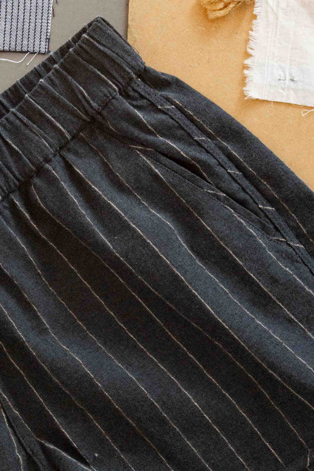 Close-up of Bridge & Burn’s Luca Track Short in Charcoal Stripe: black summer shorts with thin white vertical stripes, elastic waistband, and side pocket, shown on a brown surface near fabric swatches.