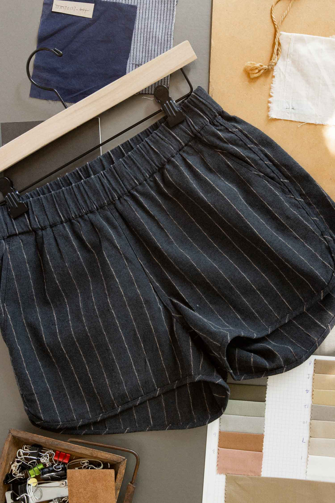 The Bridge & Burn Luca Track Short in Charcoal Stripe hangs on a black hanger, surrounded by fabric swatches, paper, and small office supplies on a tabletop.