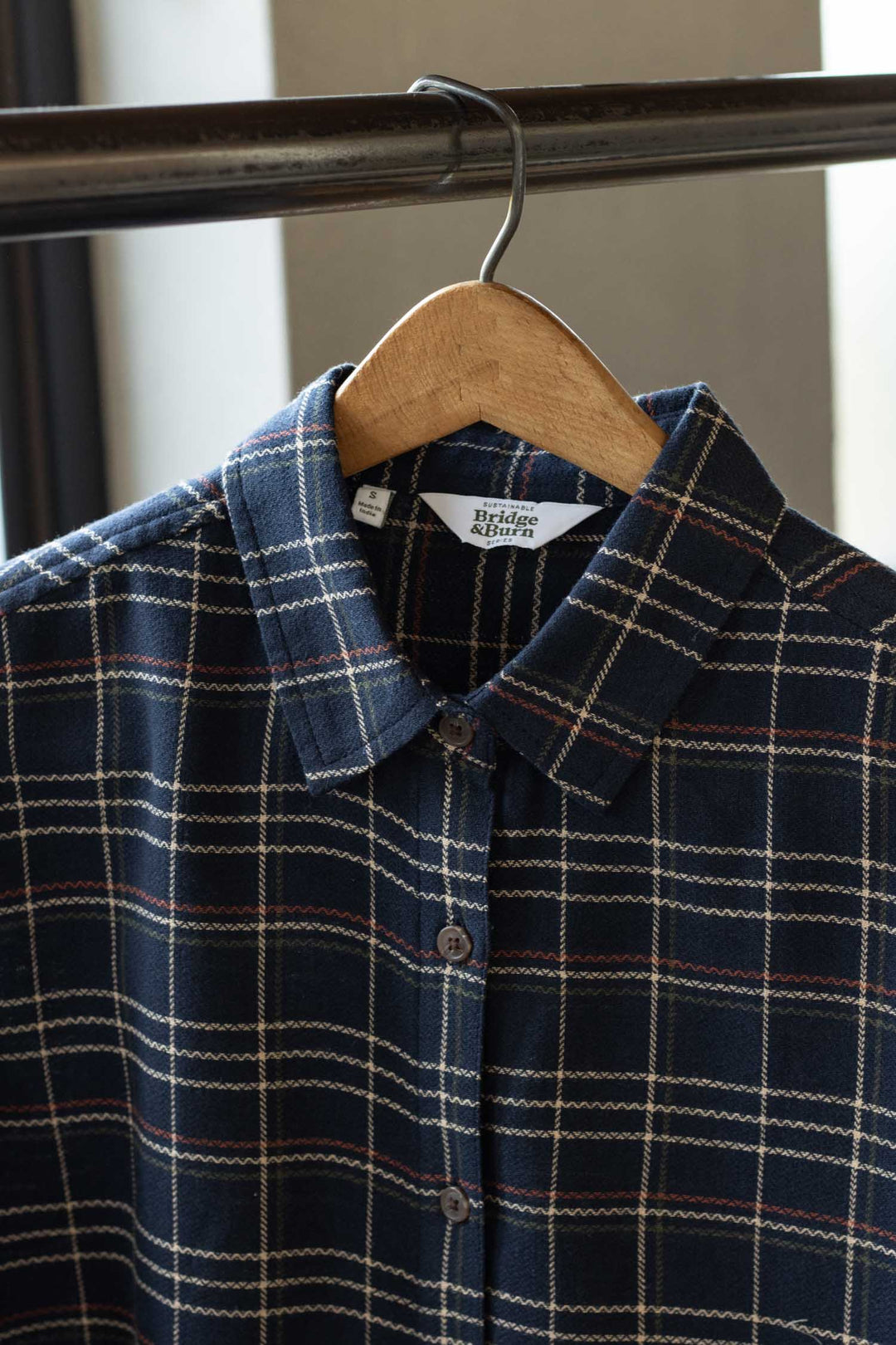 The Olivia Slim Shirt in Dusk Herringbone Plaid by Bridge & Burn, made from recycled cotton, features a navy blue pattern with white and red lines and is displayed on a wooden hanger on a metal clothing rack.