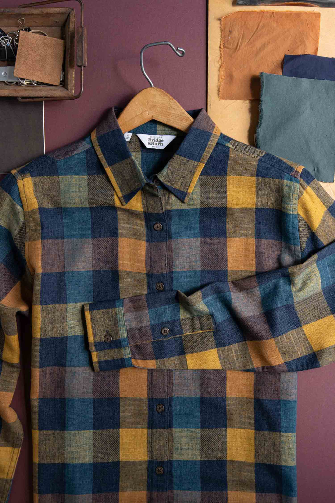 The Olivia Slim Shirt / Moonbow Plaid by Bridge & Burn—a modern, slim-fit button-up in blue, yellow, and brown plaid—hangs on a wooden hanger amid fabric swatches and sewing supplies. Made from recycled cotton.