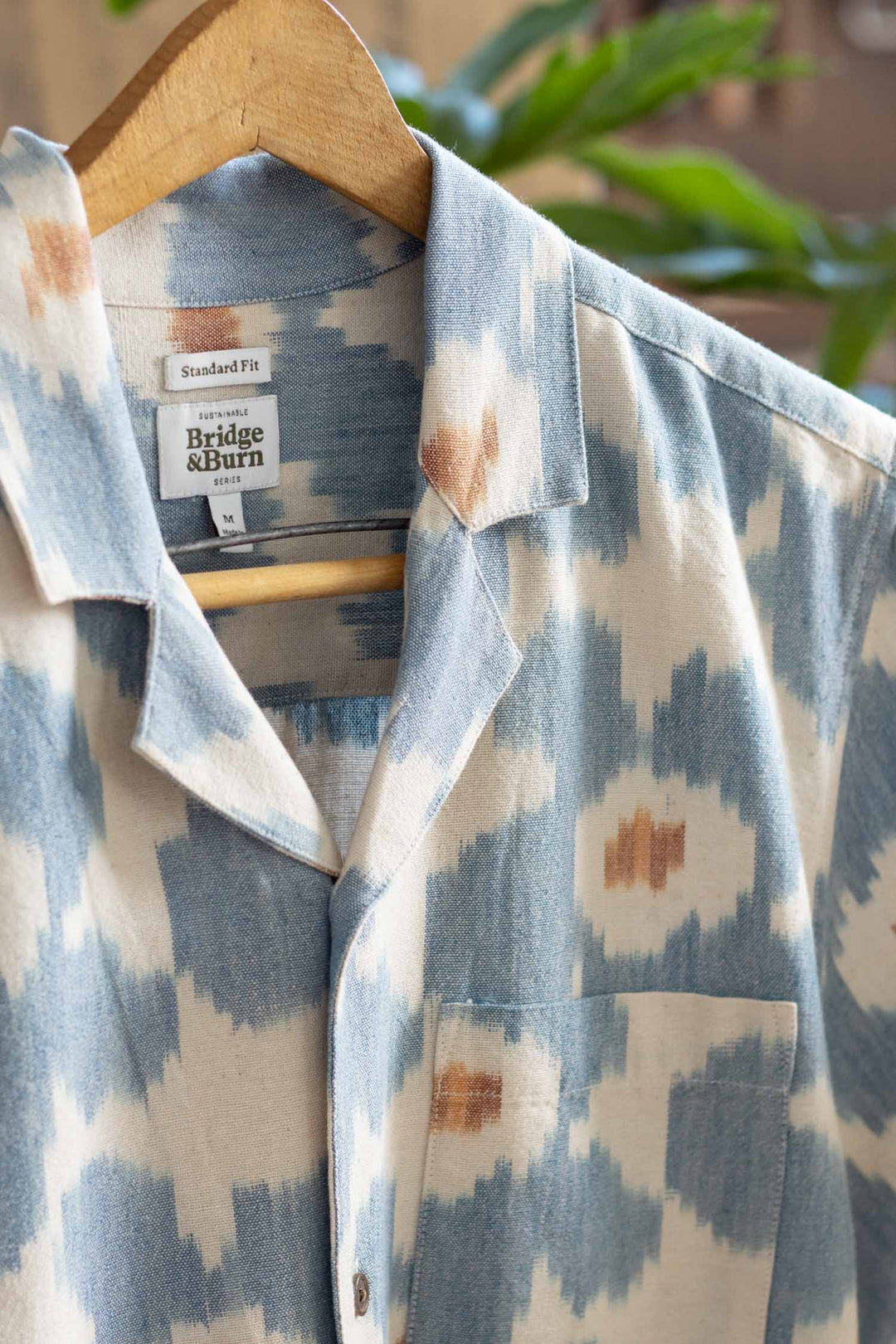 A close-up of the Bridge & Burn Bodie Shirt in Polar Blossom, featuring a blue and white pattern and retro camp collar, hangs on a wooden hanger with the label visible. Blurred green leaves appear in the background.