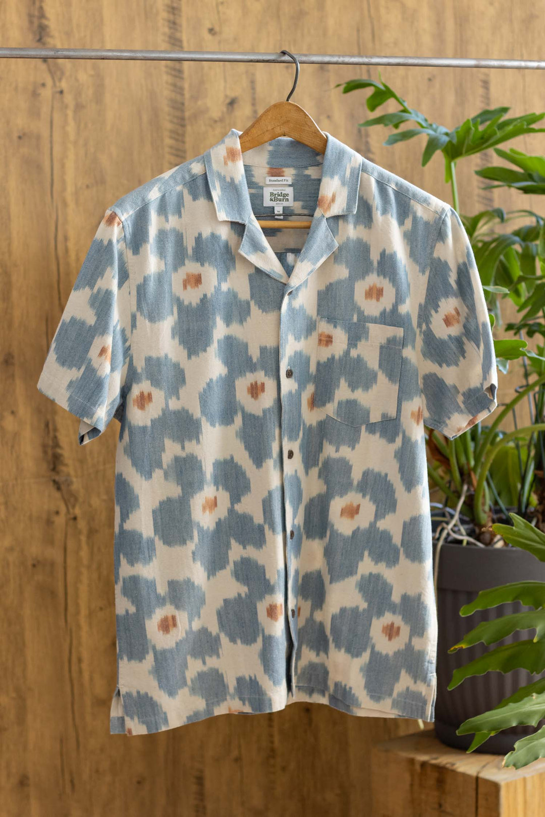 The Bodie Shirt / Polar Blossom by Bridge & Burn is a relaxed fit, short-sleeve cotton button-up featuring a blue and beige abstract floral pattern and a retro camp collar. Displayed on a hanger against wood and green plants.