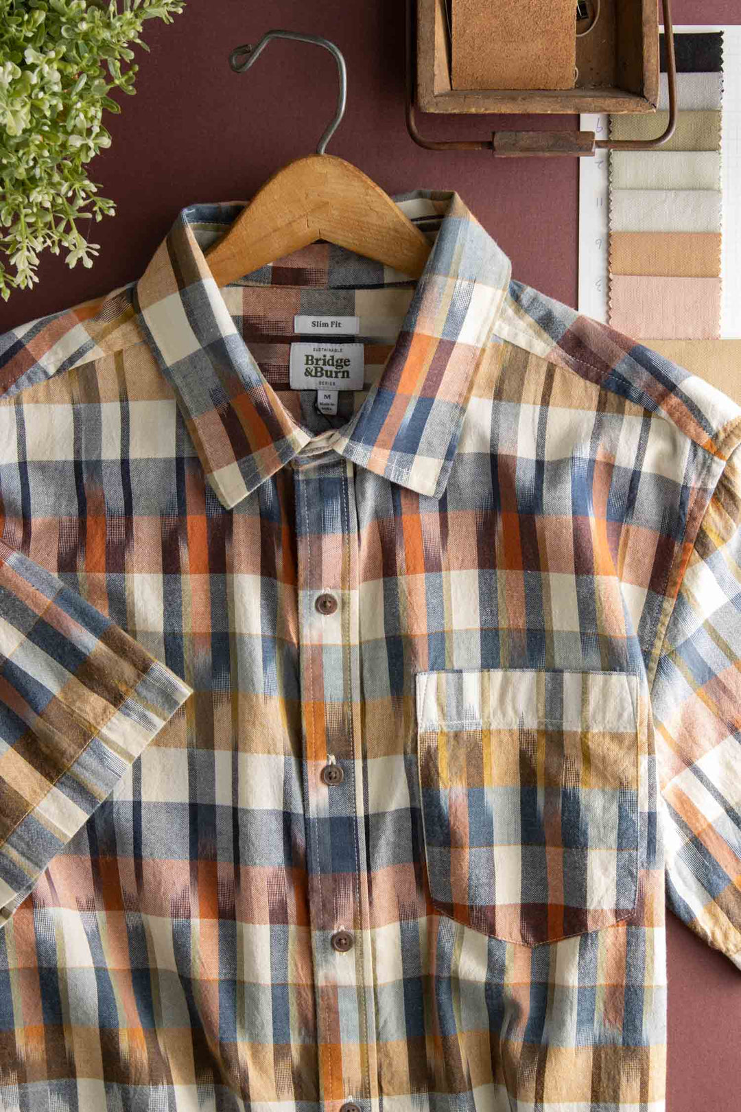 The Bridge & Burn Harbor Slim Shirt in Aurora Space Dye Plaid—featuring short sleeves, button-down front, and blue, brown, and cream plaid cotton—hangs on a wooden hanger beside fabric swatches, a plant, and a box of rolled fabric.