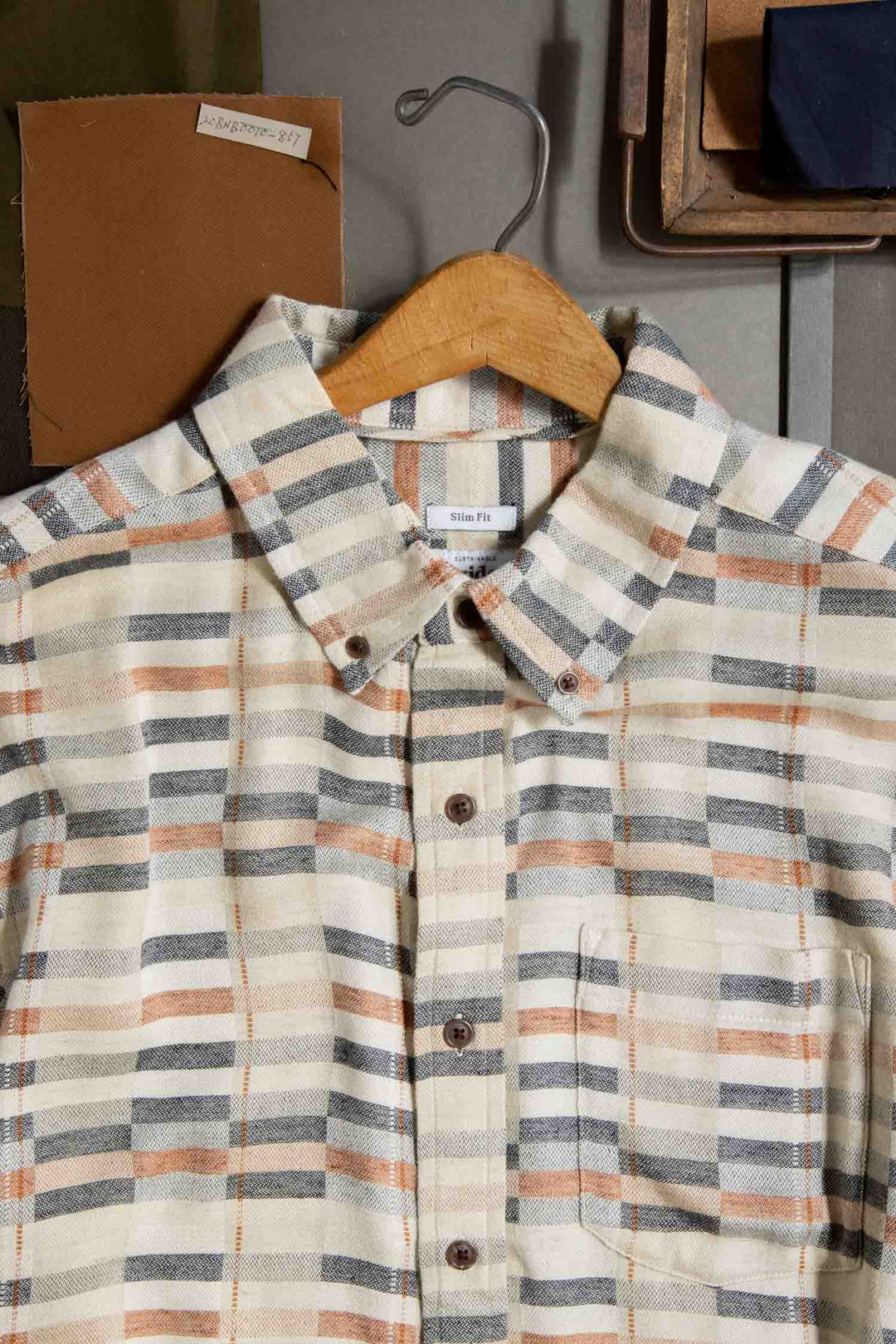 The Bridge & Burn Jordan Slim Shirt in Broadway Stripe, a modern tailored plaid with beige, gray, and orange stripes, hangs on a wooden hanger beside fabric swatches and a wooden box atop a gray surface.