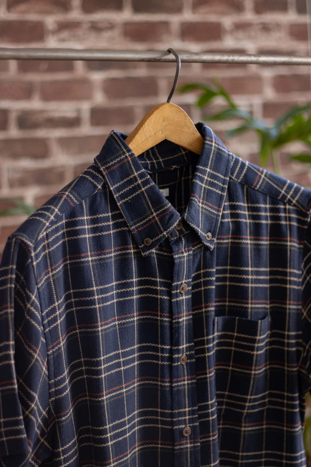 The Bridge & Burn Jordan Slim Shirt in Dusk Herringbone Plaid, featuring modern navy blue with white, beige, and red lines, hangs on a wooden hanger against a blurred brick wall background.