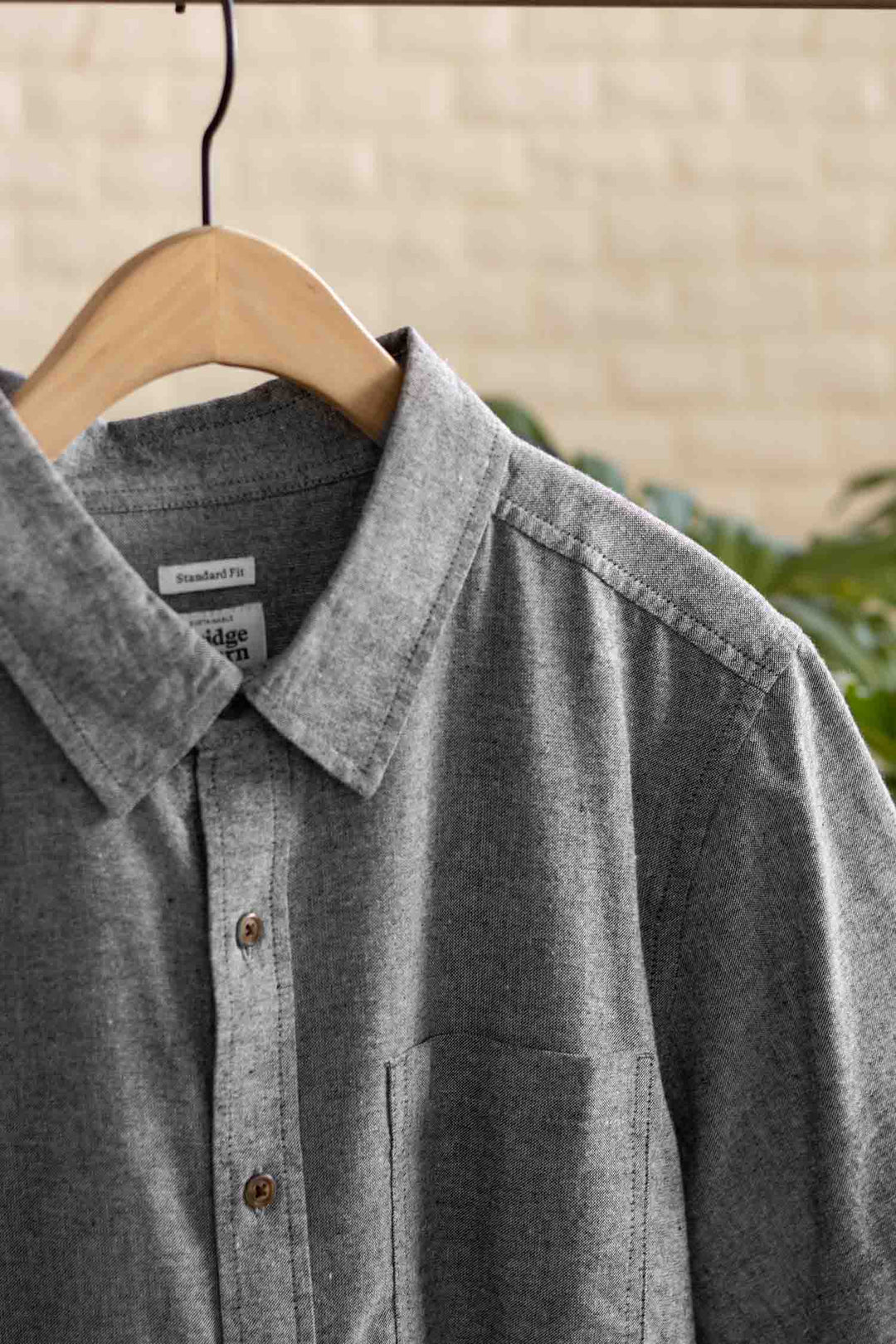 The Bridge & Burn Larson Shirt in Iron Chambray, a classic fit men's button-up with chest pocket, made from recycled cotton, hangs on a wooden hanger against softly blurred greenery and a beige wall.