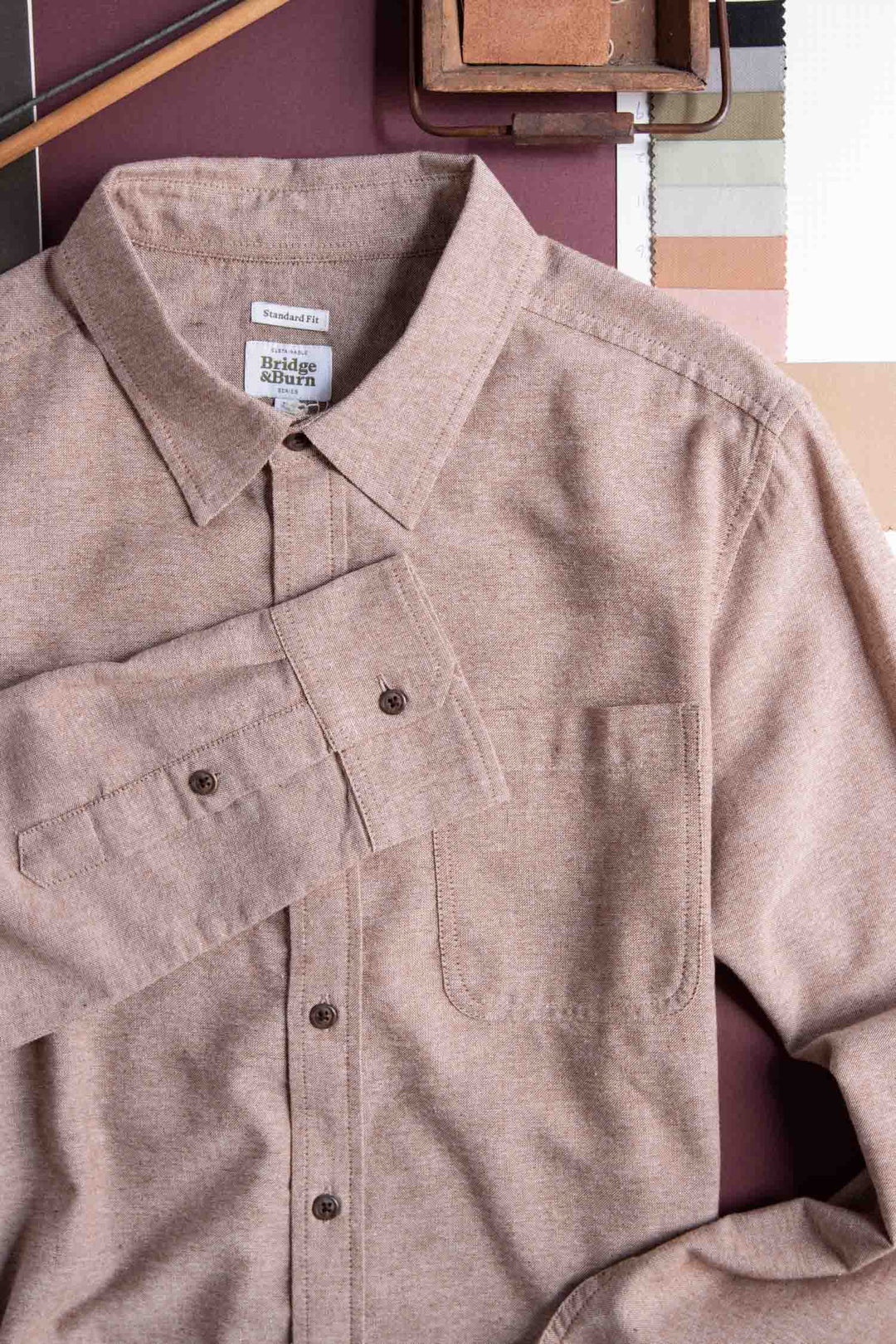 The Bridge & Burn Larson Shirt in Rust Chambray, a classic fit men's button-up made from recycled cotton, is displayed next to a color palette and sketching tools, evoking a fashion or design setting.