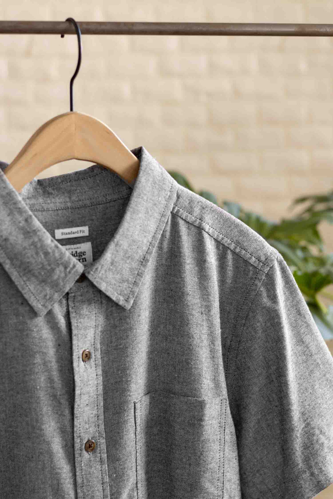 The Malcolm Shirt in Iron Chambray by Bridge & Burn—a gray recycled cotton short sleeve button-up with brown buttons and a chest pocket—hangs on a wooden hanger, set against green plants and a beige brick wall.