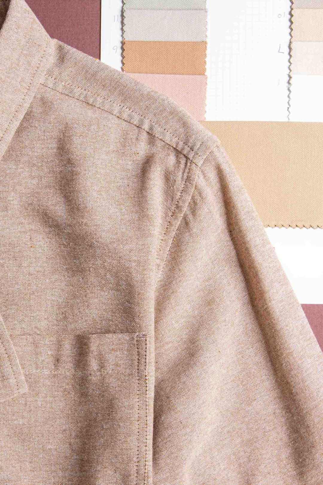 A close-up of the Malcolm Shirt in Rust Chambray by Bridge & Burn shows its shoulder and chest pocket, with neutral-toned fabric swatches in the background, highlighting its eco-friendly design.