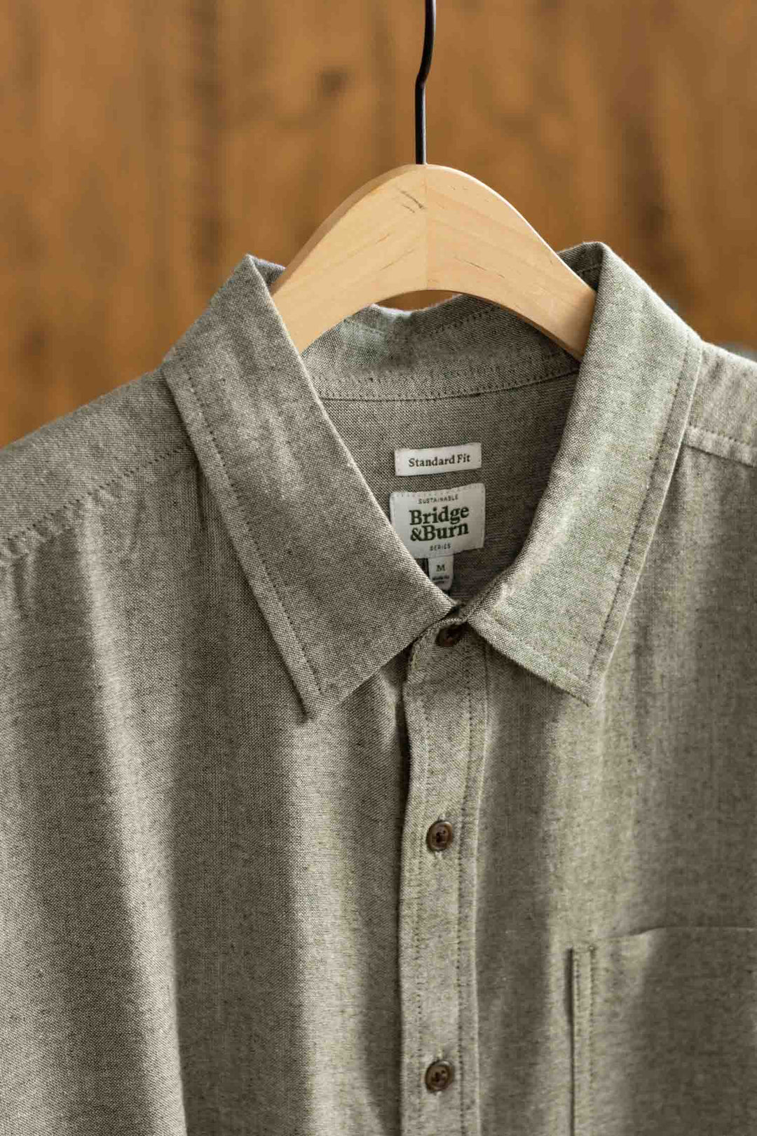 The Malcolm Shirt in Sage Chambray by Bridge & Burn hangs on a wooden hanger. This sustainable men’s short sleeve button-up features a collar, chest pocket, and Standard Fit label, displayed against a wooden background.