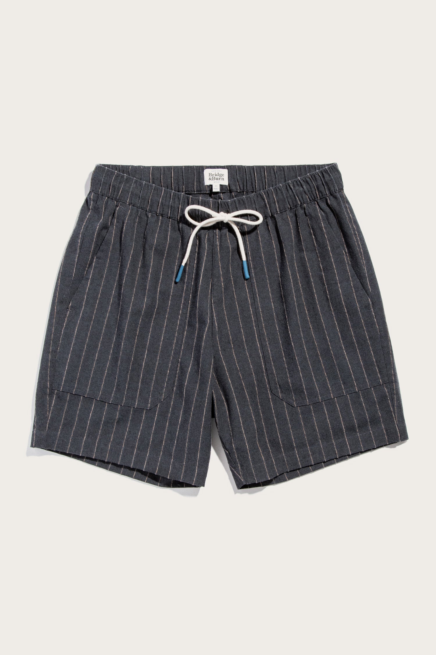The Noah Short by Bridge & Burn in Charcoal Stripe features white vertical pinstripes, an elastic waist with a blue-tipped white drawstring, and two front pockets. Shown laid flat on a light background.