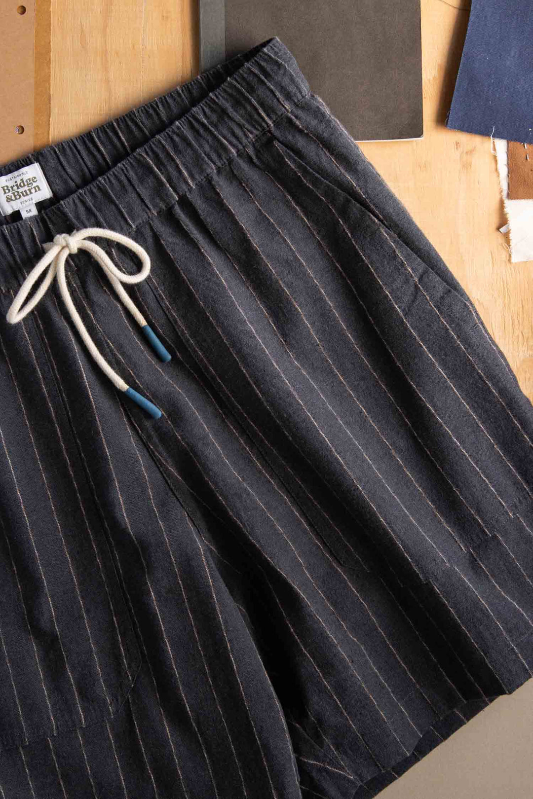 The Bridge & Burn Noah Short in Charcoal Stripe features relaxed pull-on styling, an elastic waist, and a drawstring, shown laid flat on a wooden surface with fabric swatches in the background.