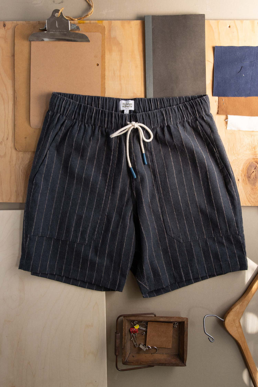 The Bridge & Burn Noah Short in Charcoal Stripe with an elastic waist and white drawstrings is laid flat on a wooden surface, surrounded by clipboards, papers, a tray with keys, and other office supplies.