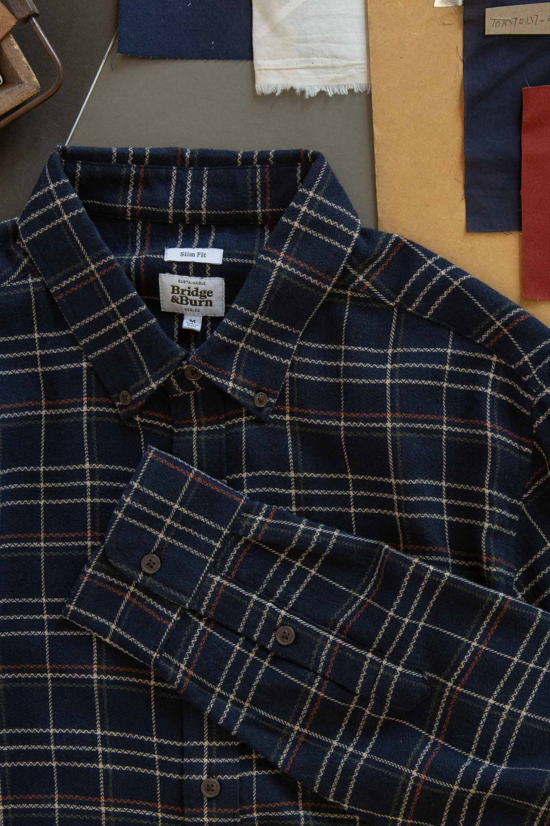 The Bridge & Burn Sutton Slim Shirt in Dusk Herringbone Plaid features a navy blue plaid with beige and red lines, shown flat with fabric swatches and pattern papers, highlighting its minimalist design and refined craftsmanship.