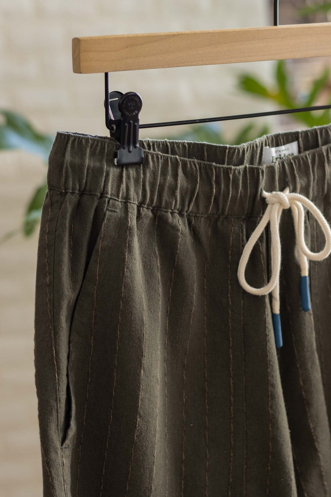 Close-up of Bridge & Burn’s Noah Short in Olive Stripe, featuring a relaxed pull-on fit, white ties, subtle vertical stitching, and cotton-linen blend fabric. Shorts hang from a wooden hanger with blurred greenery in the background.