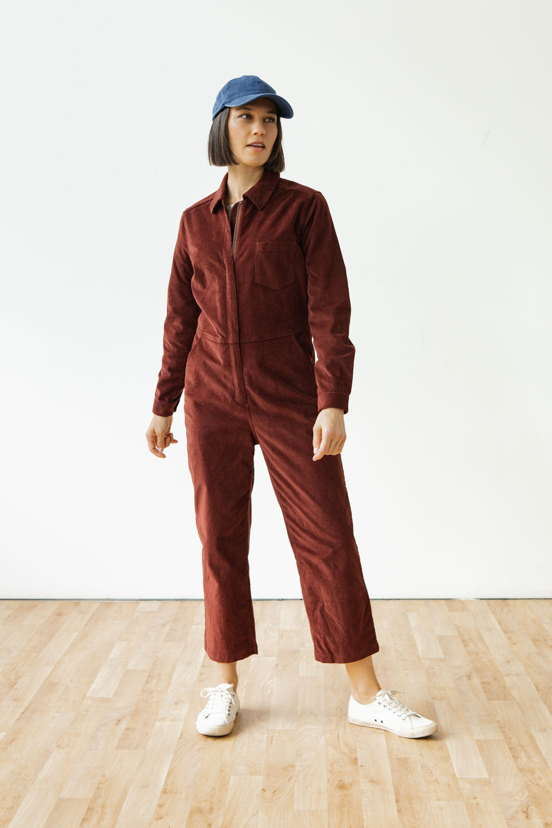 A person in a blue cap, Bridge & Burn Faye Jumpsuit in Dark Red Pinwale, and white sneakers stands on a light wooden floor against a plain white background.