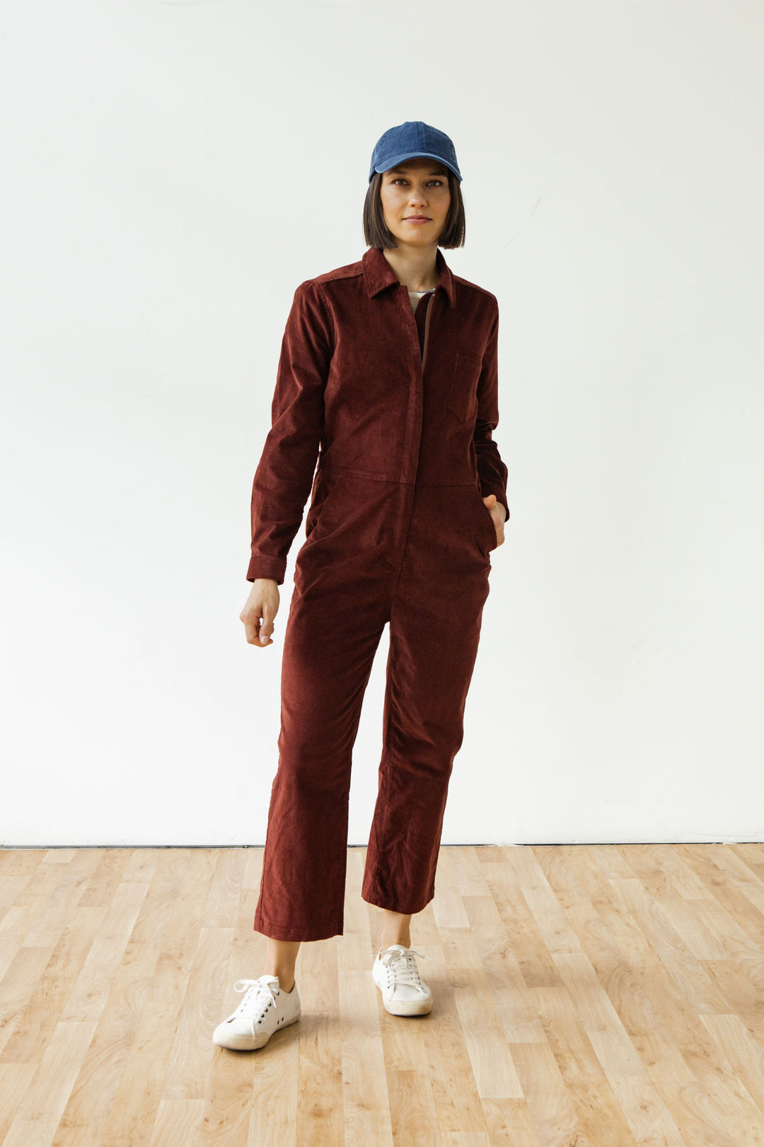 A person stands indoors on a wooden floor, wearing the Bridge & Burn Faye Jumpsuit in Dark Red Pinwale. They have one hand in a pocket, wear white sneakers and a blue cap, with a neutral expression against a white wall.