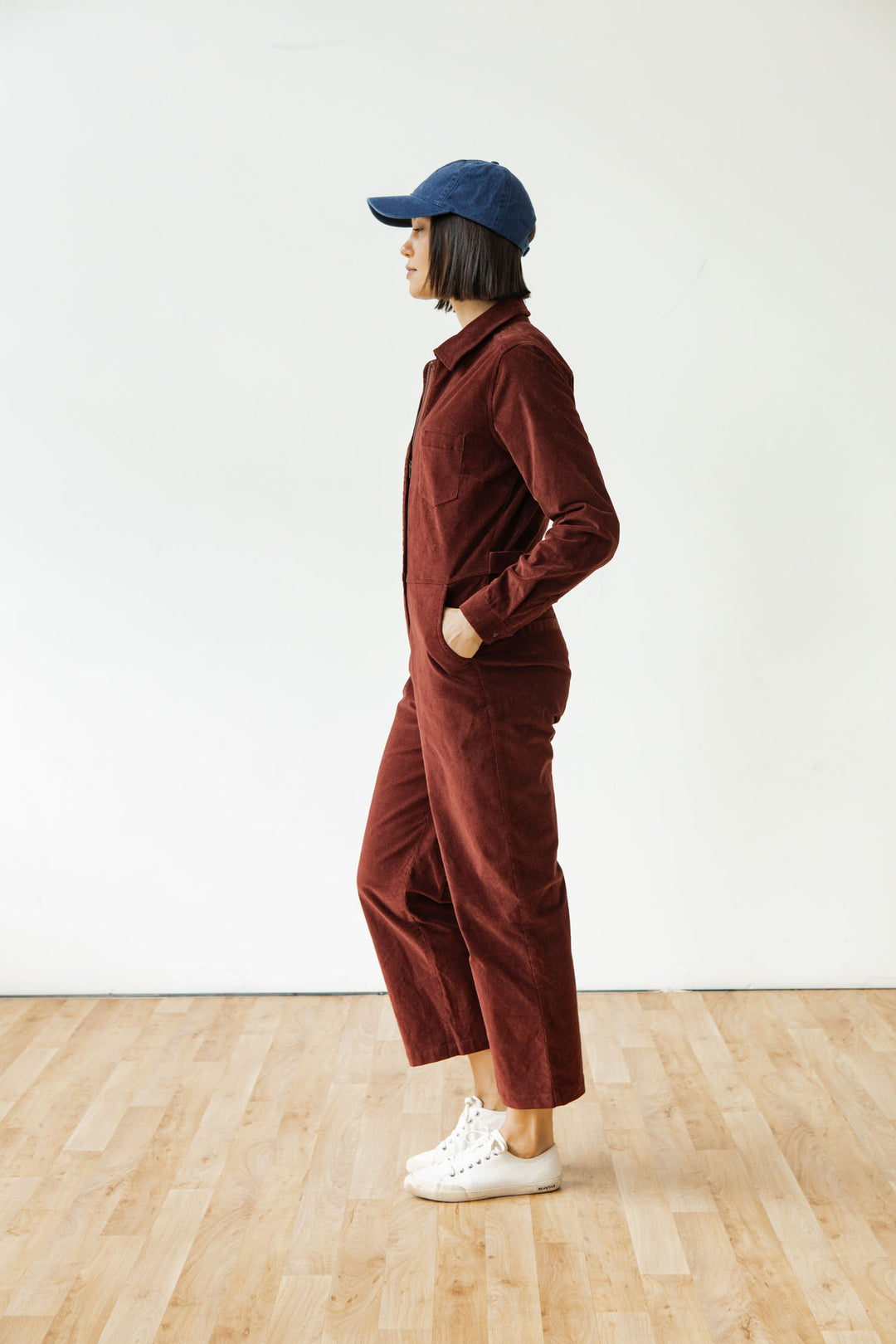 A person wears the Bridge & Burn Faye Jumpsuit in Dark Red Pinwale, paired with white sneakers and a blue cap, standing indoors on a wooden floor against a plain white wall, facing left with hands in pockets.