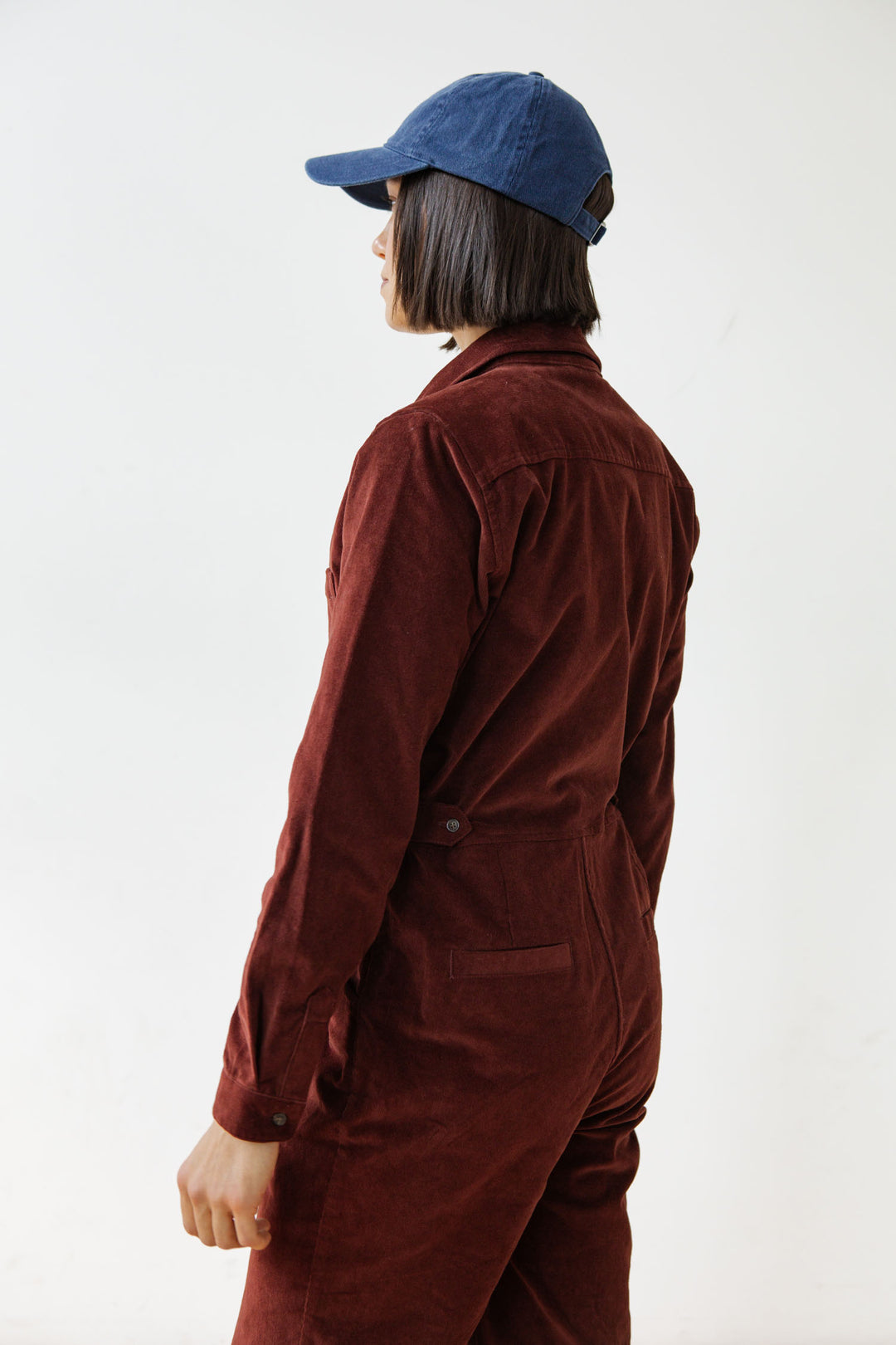 A person with short dark hair, wearing a blue baseball cap and the Bridge & Burn Faye Jumpsuit in Dark Red Pinwale, stands facing away from the camera against a plain light background.
